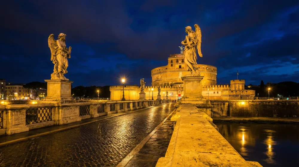 Illuminated marble angel statues on Ponte Sant'Angelo with the glowing fortress in the background at night