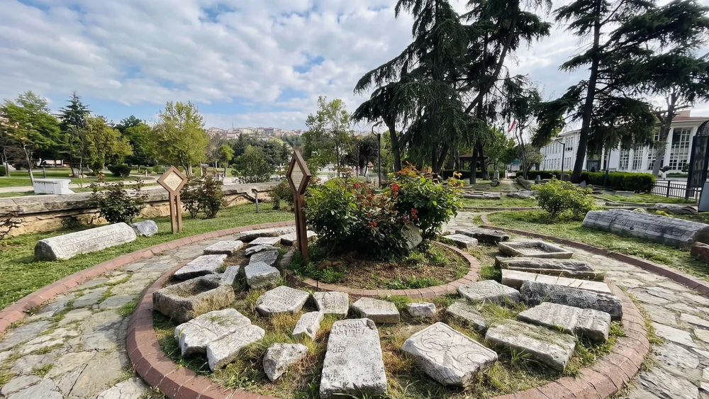 Parke zemin ve çimenlik üzerinde dağınık duran oyma taş parçaları ve mezar taşı bölümleri; çevrede ahşap bilgi tabelaları ve ağaçlar var.