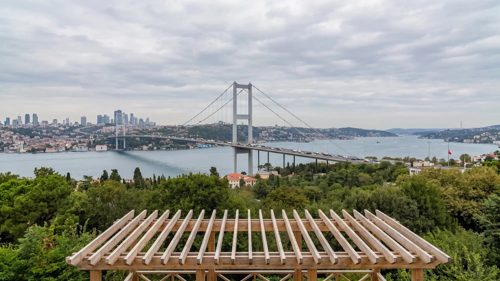 Ahşap korkuluklu bir terastan ağaçların üzerinden görünen Boğaz köprüsü ve Boğaz