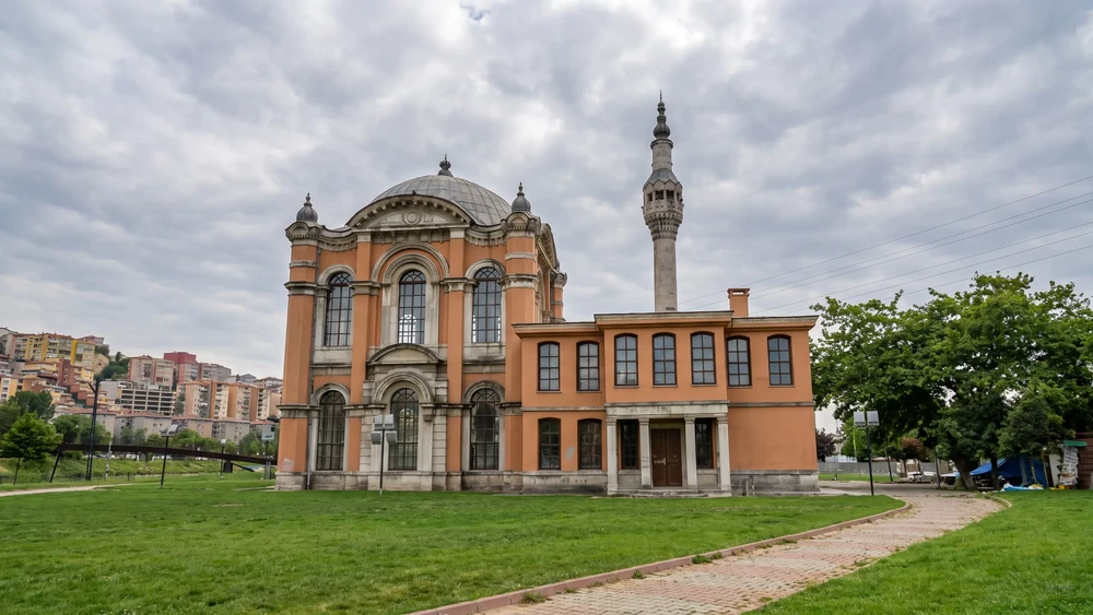 Çimenlik bir alanın karşısında, kubbeli ve tek minareli, kiremit kırmızısı duvarlı cami cephesi.