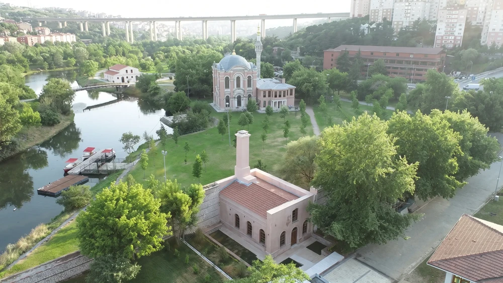 Havadan görünüm: dere kıyısında pembe duvarlı, bacalı bir bina; ileride çimenlik alanın ucunda kubbeli ve tek minareli bir cami.