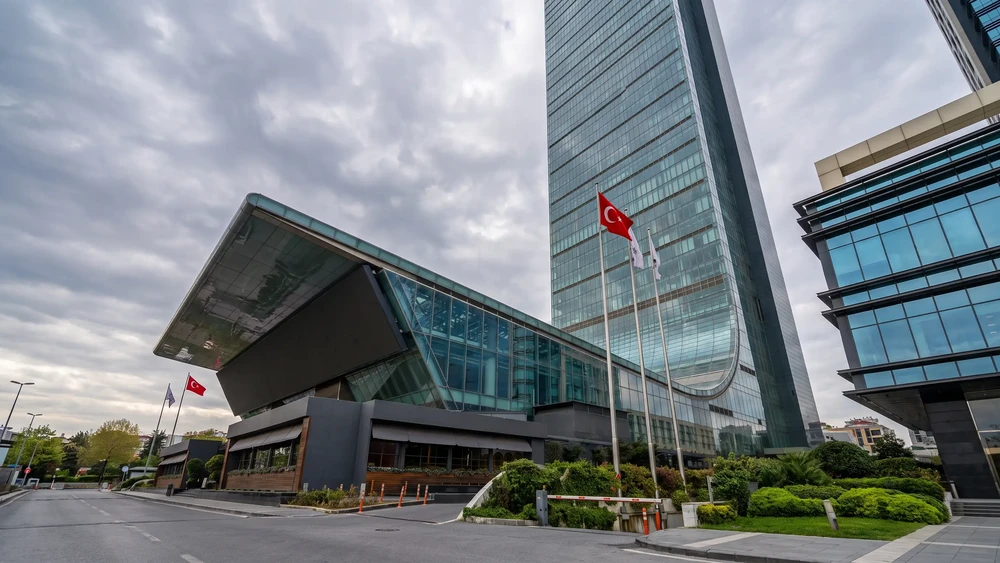 Cam cepheli yüksek Sapphire kulesi ve yanındaki eğimli çatılı alçak yapı, önde bayraklar