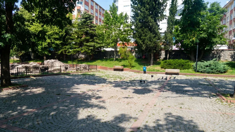 Parkta taş döşemeli açık alan, banklar ve ağaçlar