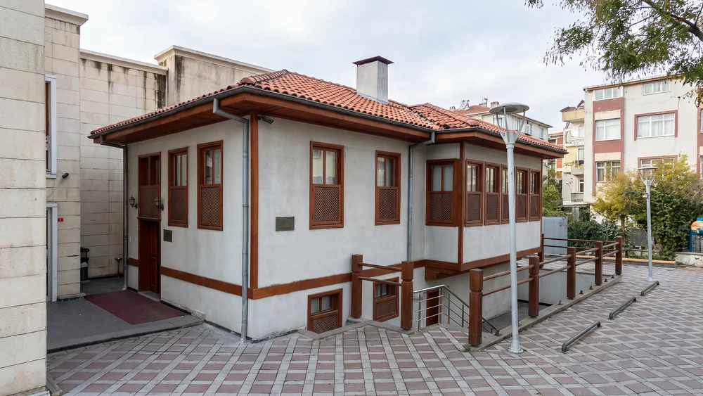 Ahşap saçaklı, beyaz cepheli ve kahverengi panjurlu iki katlı selâmlık tarzı bina, önünde taş döşeli meydan